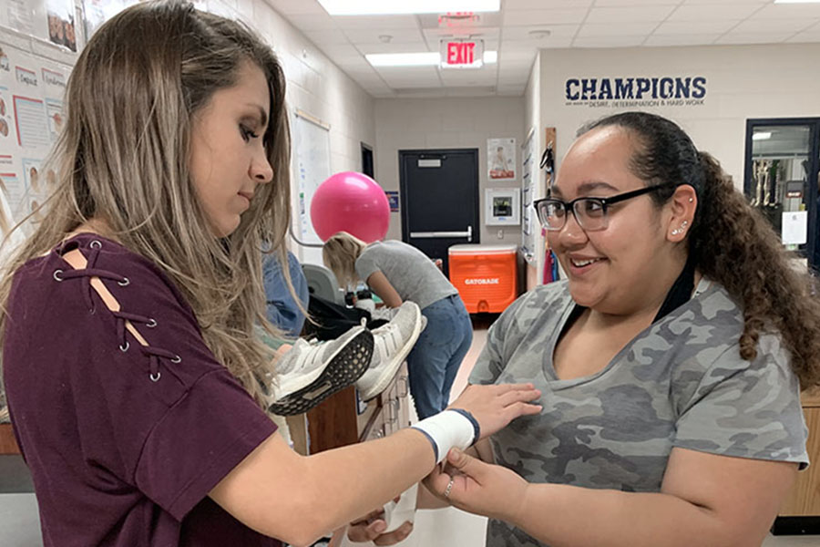 With a smile on her face, junior Leila Martinez, tapes junior Lexi Stephens’s wrist for extra practice . “I was practicing my taping in case an athlete came in and needed to be taped,” Martinez said. Trainers constantly practice taping each other to ensure their skills are always as best as they can be. 